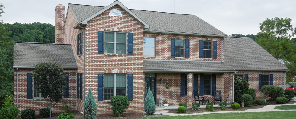 A large two-story brick house with a gray shingled roof and navy blue shutters on the windows. The house, by Steve L. Dellinger Contractor, features a front porch with columns, various bushes and small trees in the landscaped yard, and a curved walkway leading to the front steps.