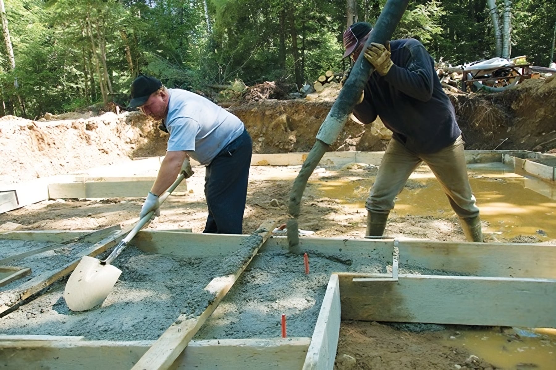 Two construction workers, Steve L. Dellinger Contractor and his partner, are pouring and spreading flat concrete into wooden molds outdoors. One worker uses a shovel to level the concrete while Steve directs the flow from a hose. The surroundings include trees and construction materials.