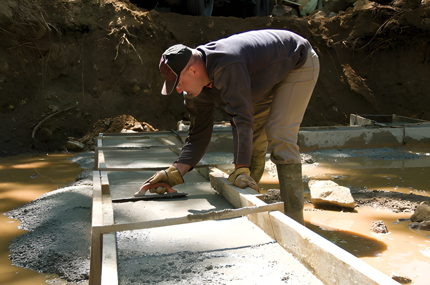 Steve L. Dellinger Contractor, wearing work clothes, a cap, and gloves, is bent over a wooden framework being constructed in a muddy area. They appear to be using a tool to smooth a concrete mixture within the framework. Sunlight filters through the trees, casting shadows on the workspace for this residential and commercial project.