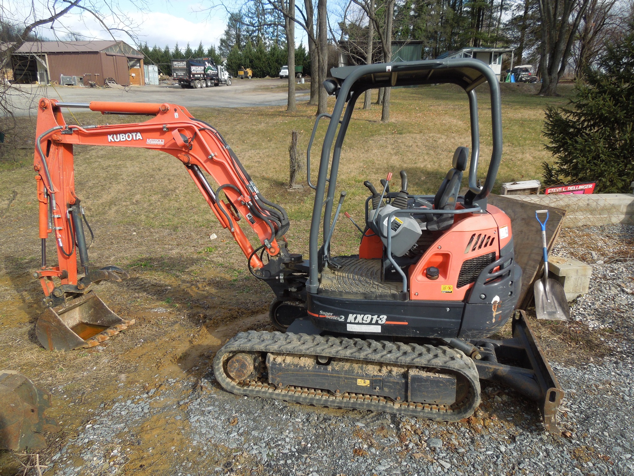 A red Kubota KX91-3 mini excavator, belonging to Steve L. Dellinger Contractor, is parked on a gravel surface. The excavator features a small front loader bucket and an operator's seat enclosed by a roll cage. Trees, a building, and a parked car are visible in the background—ideal for residential and commercial excavating projects.