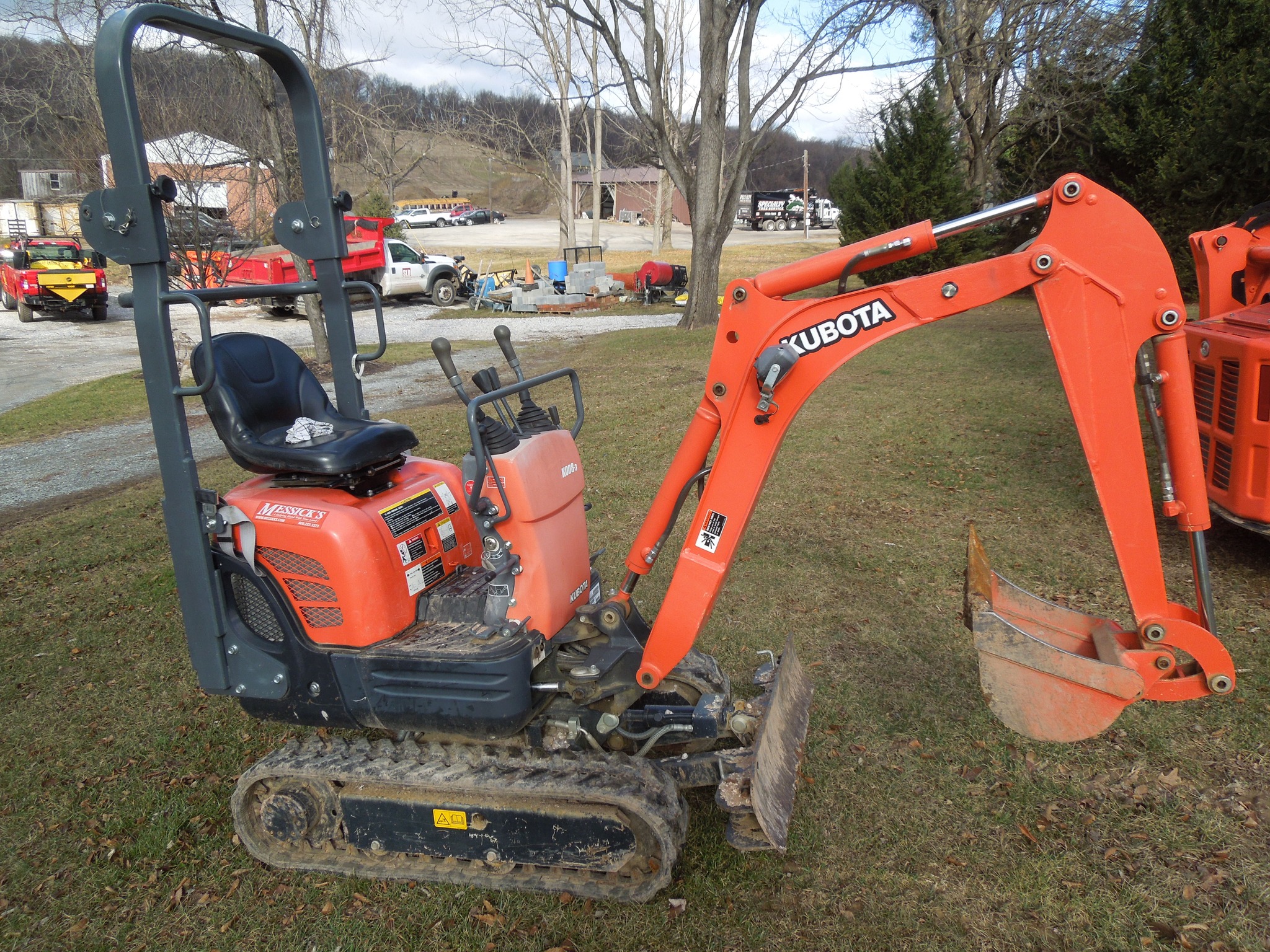 A small orange Kubota excavator is parked on a grassy area. It features a seated operating area and a digging arm with a bucket attachment, ideal for residential and commercial excavating projects. Various vehicles and equipment are visible in the background, with trees and buildings nearby.