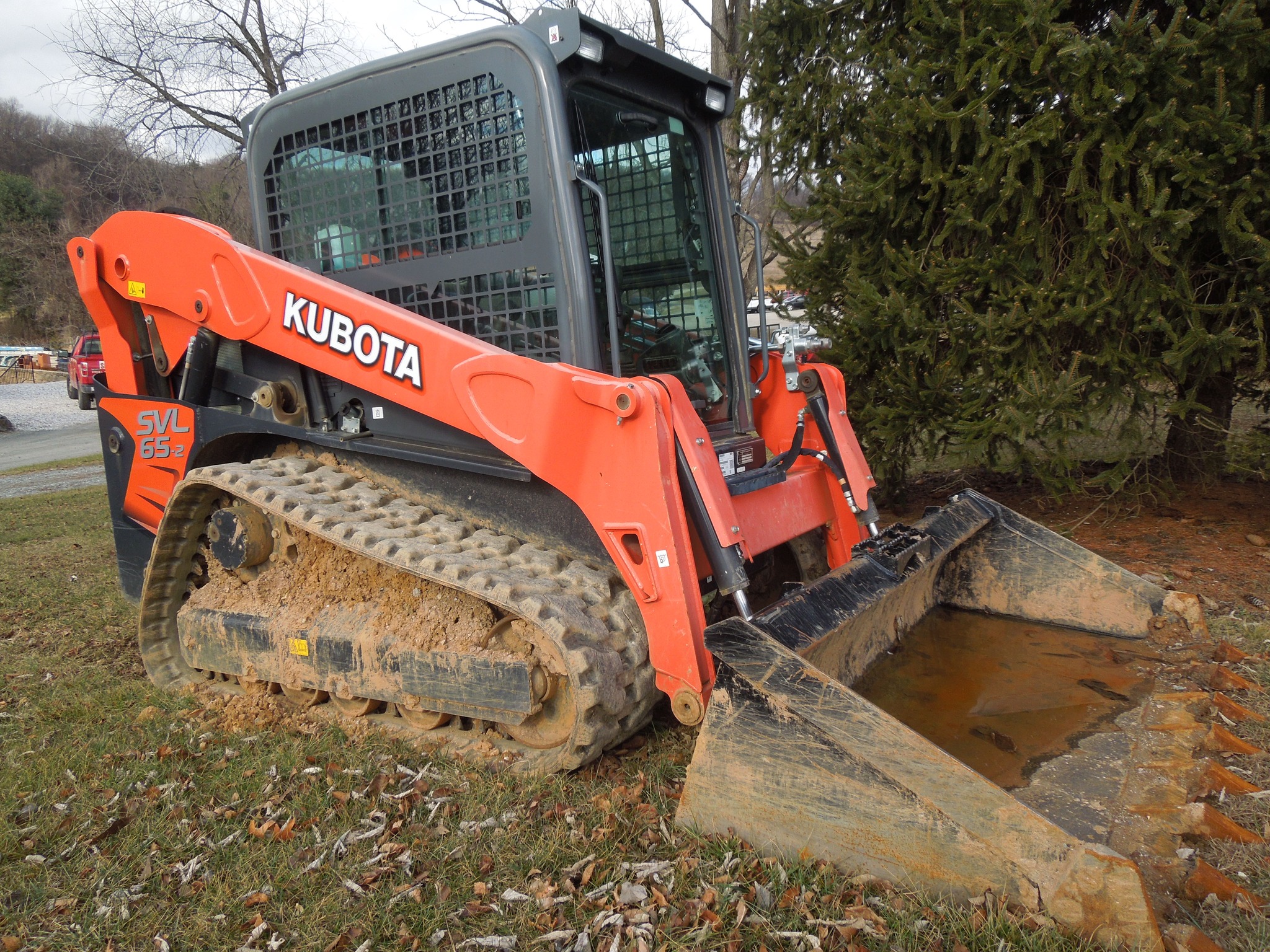 A red Kubota skid steer loader with muddy tracks and a filled bucket is parked on a grassy area. The loader, suitable for residential and commercial use, has a protective cab, and trees are visible in the background.