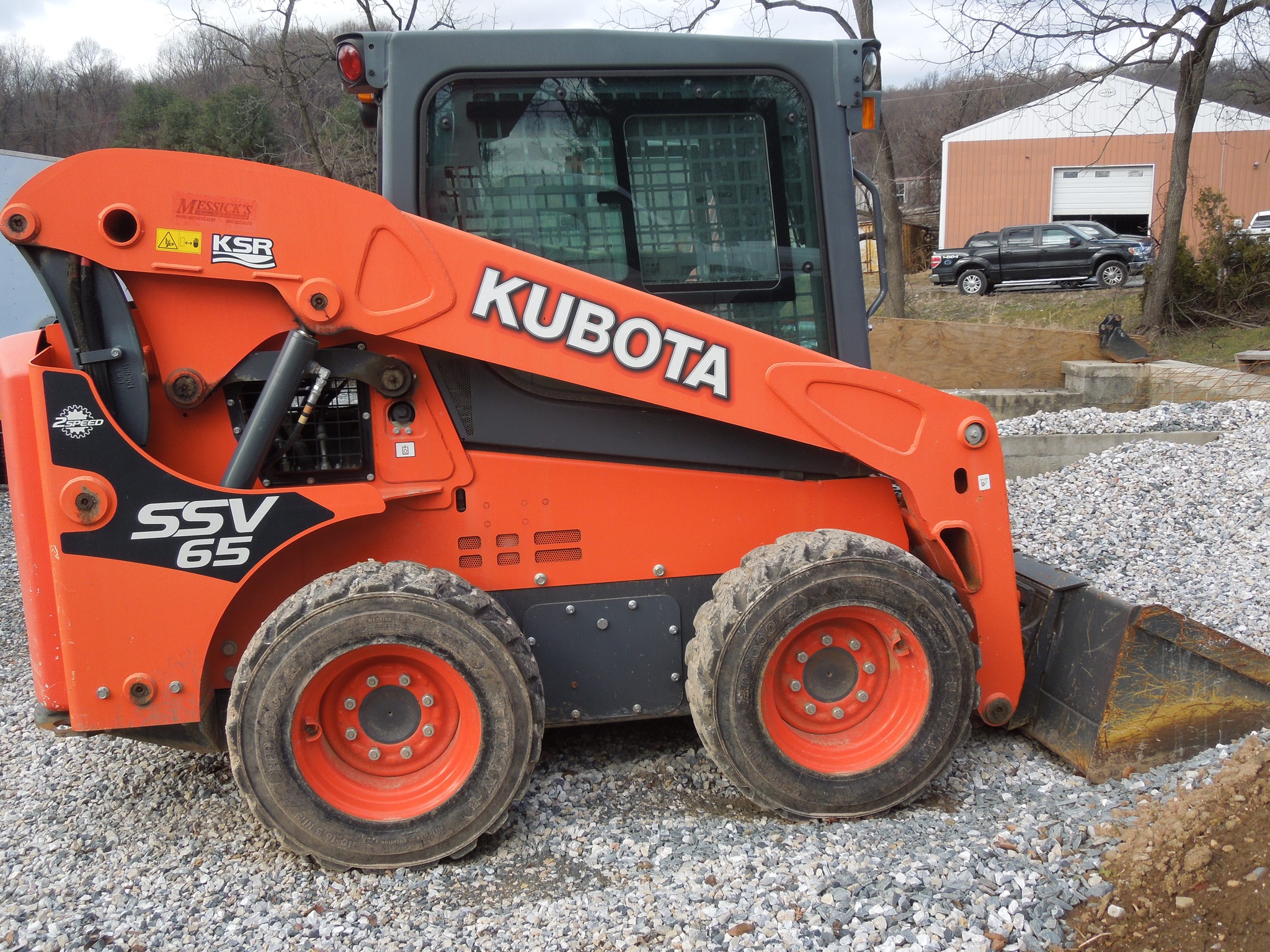 An orange Kubota SSV65 skid steer loader is parked on a gravel surface outdoors. The machinery, with large tires and a front bucket attachment, belongs to Steve L. Dellinger Contractor. In the background, there are buildings, trees, and a hill. Ideal for residential and commercial excavating tasks.