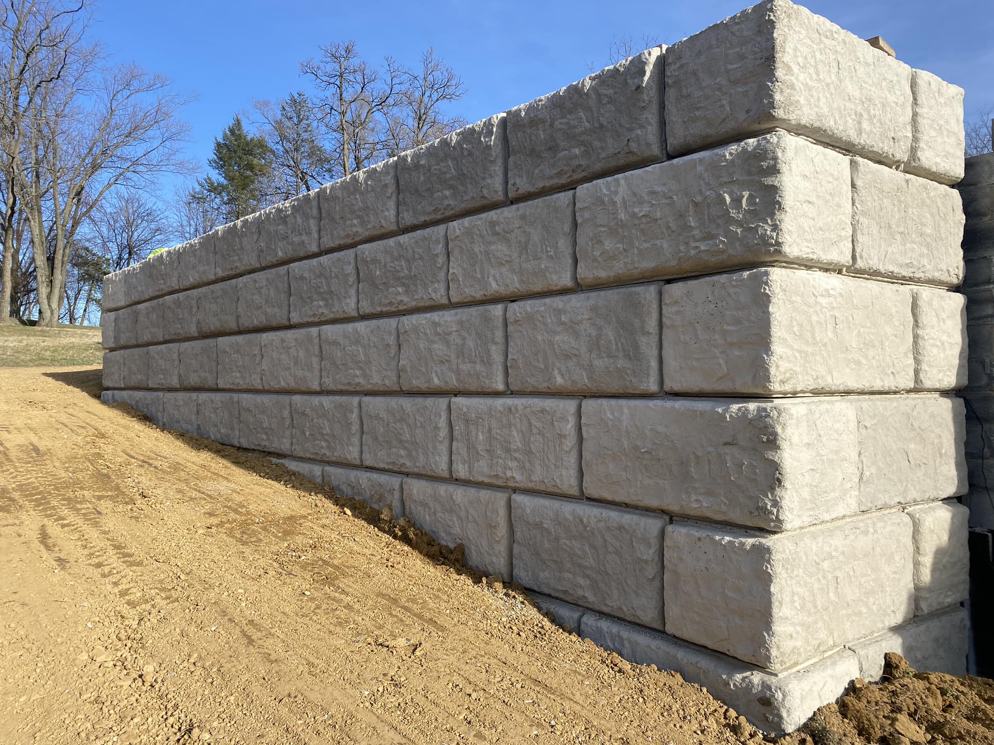 A large stone retaining wall made up of rectangular concrete blocks with a rough surface is seen along a slope of soil. The ground is barren, and leafless trees are visible in the background under a clear blue sky, reflecting the precision of Steve L. Dellinger Contractor’s work in residential and commercial excavating.