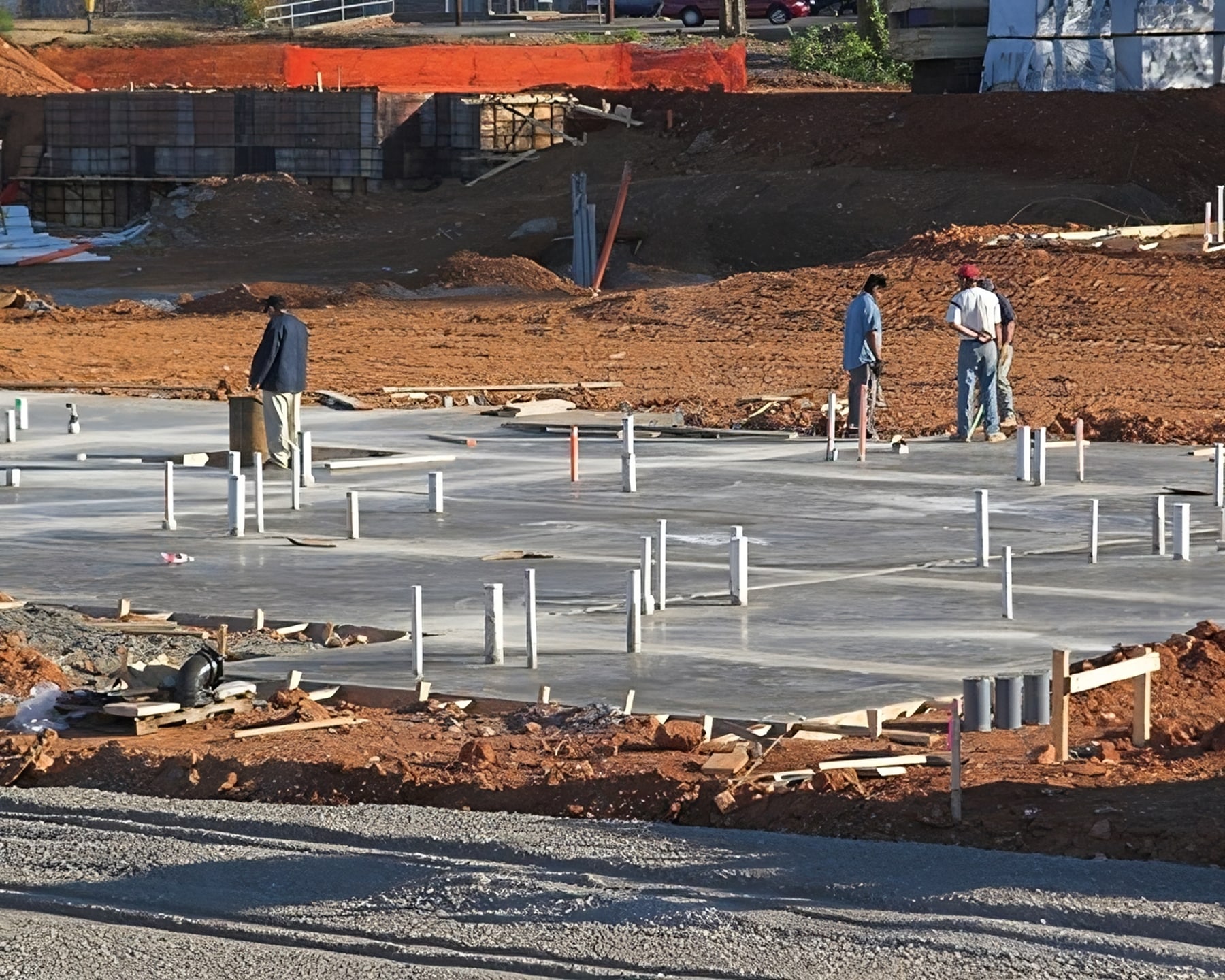 Three construction workers are standing on a flat concrete slab at a construction site, where numerous white utility pipes protrude. The site, overseen by Steve L. Dellinger Contractor, is surrounded by dirt and construction materials with an orange safety barrier in the background.