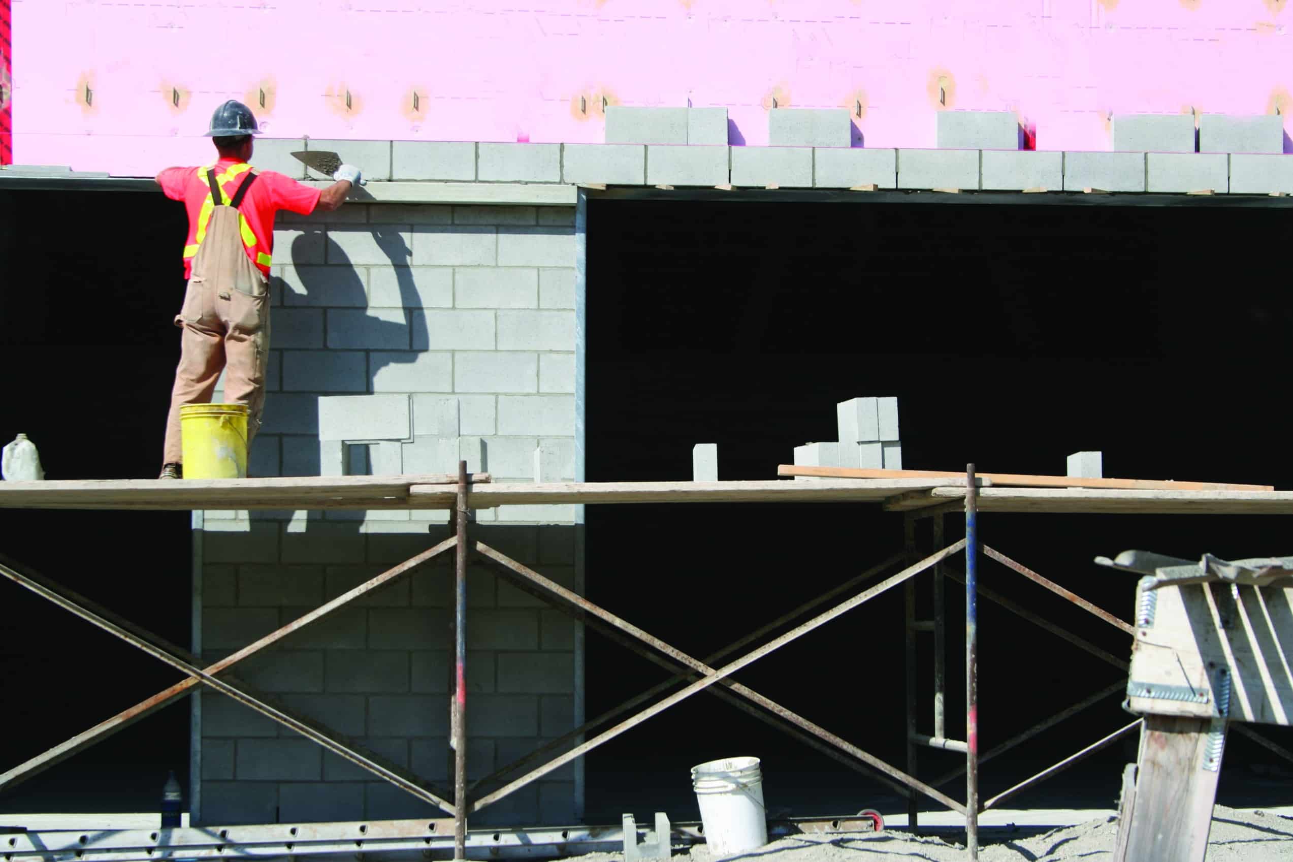 A construction worker wearing a red shirt, yellow safety vest, and hard hat stands on scaffolding, building a wall with gray concrete blocks. The building wall has visible insulation in the upper section. Another bucket and construction materials from Steve L. Dellinger Contractor are on the ground below.