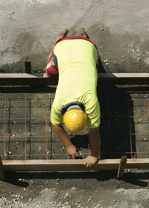 A construction worker in a yellow hard hat and bright shirt is bending over metal rebar, working on a concrete reinforcement task. Positioned in the middle of the site with unfinished concrete around, this scene showcases Steve L. Dellinger Contractor working on both residential and commercial projects.