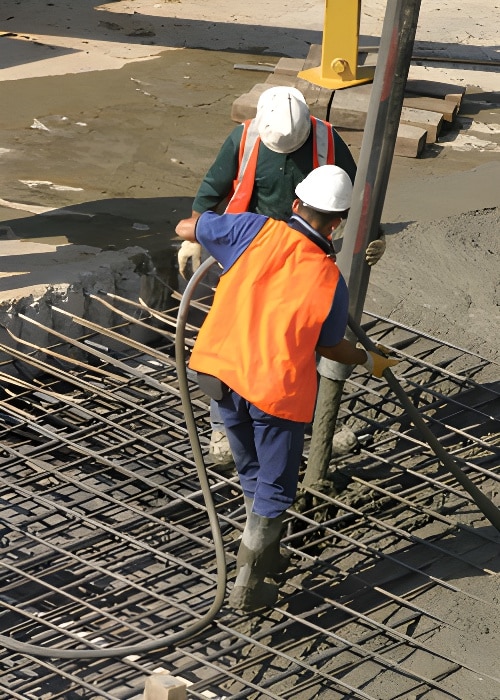 Two construction workers wearing white helmets and orange safety vests are pouring flat concrete from a large hose onto a rebar grid. They are working on a construction site with wet concrete and tools visible. Both workers are focused on directing the concrete flow for residential and commercial projects.