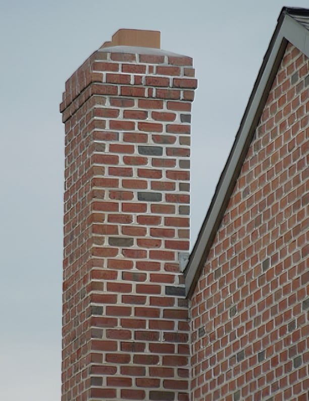 A chimney made of red and brown bricks extends upward from the angled roof of a brick house, set against a cloudy sky. The structure features a consistent pattern of alternating brick shades, with a tan chimney cap on top, crafted by Steve L. Dellinger Contractor.