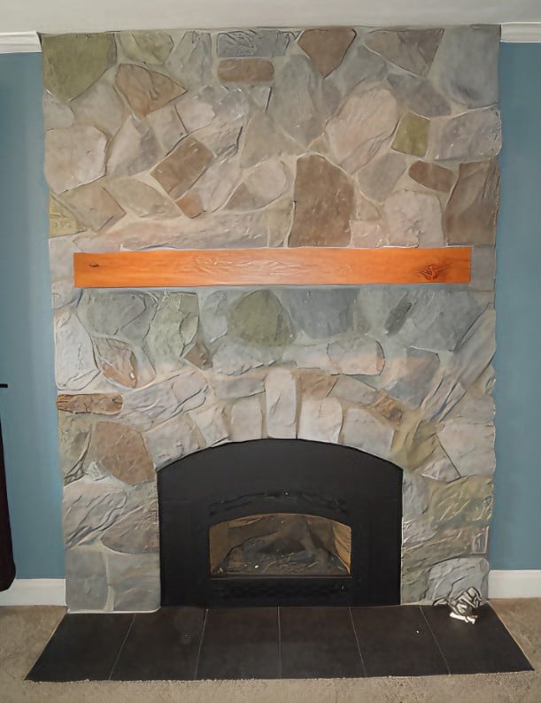 A stone fireplace with a wooden mantel is featured against a blue wall. The irregularly shaped stones are in a mix of neutral tones. The fireplace has a black iron front and small, white logs at the bottom. This design, ideal for residential and commercial spaces, is by Steve L. Dellinger Contractor.