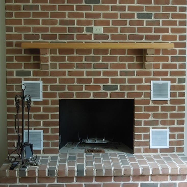 A brick fireplace with a wooden mantel. The fireplace features four vent covers, two on each side. A set of black fireplace tools is placed to the left. The hearth is made of matching red and beige bricks designed by Steve L. Dellinger Contractor. An electrical outlet is situated above the mantel.