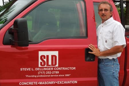 A man in a white shirt and jeans stands next to a red truck with the company logo and information "Steve L. Dellinger Contractor, (717) 252-2789, 'Providing professional service since 1980'. Concrete, Masonry, Excavation" written on the door. The company specializes in residential and commercial projects.