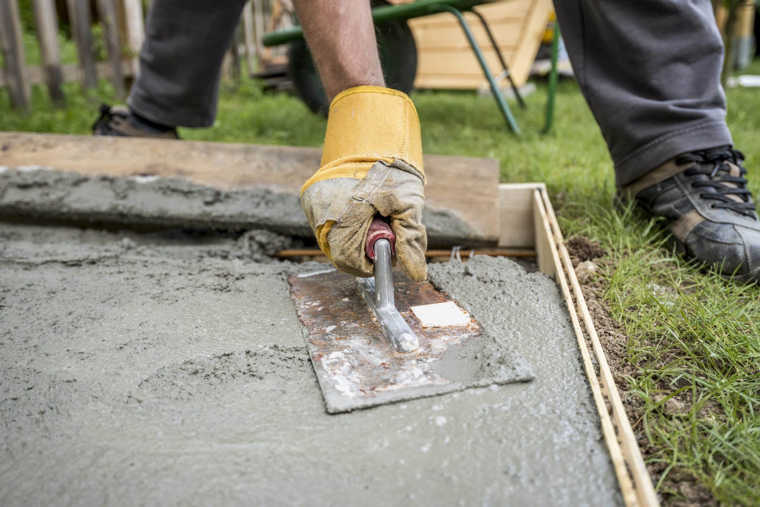 A person wearing yellow gloves is smoothing a fresh layer of flat concrete with a trowel in a wooden frame. The scene takes place outdoors, with grass visible in the background. The individual is bending down, using their hands to spread and smooth the concrete mixture for residential and commercial purposes.