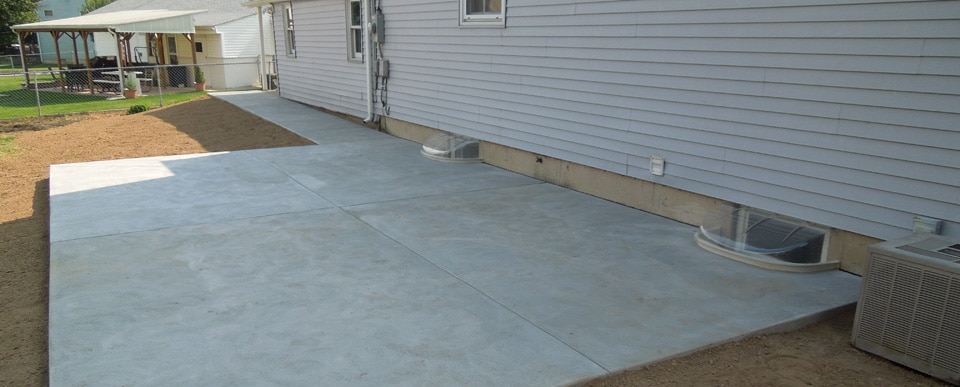 A freshly laid concrete basement walkout area runs alongside a gray house, expertly crafted by Steve L. Dellinger Contractor. Two basement window wells with plastic covers are visible along the house wall. The area is bordered by a patch of dirt, and a fenced yard with a gazebo can be seen in the background.