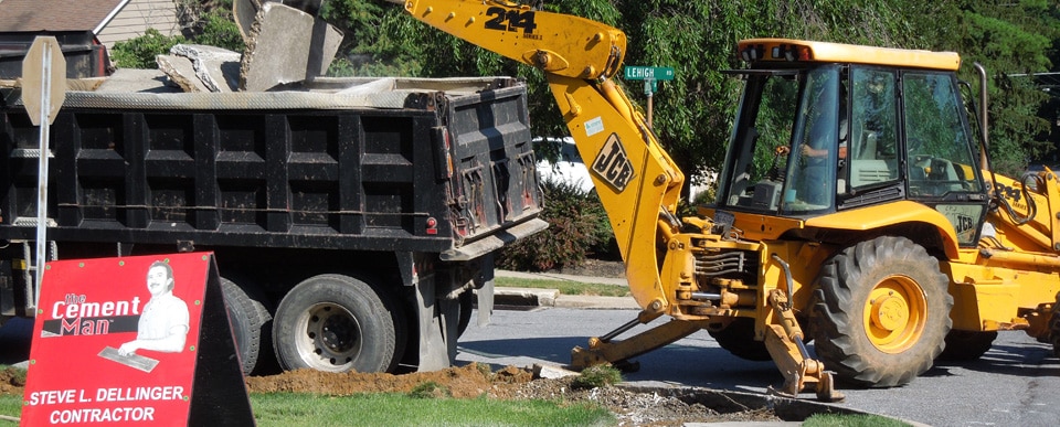 A backhoe loader dumps debris into a dump truck at a construction site on a residential street. A red sign on the ground reads "Steve L. Dellinger Contractor" with an illustration of a person. Trees and a house are visible in the background, highlighting their expertise in both residential and commercial excavating projects.