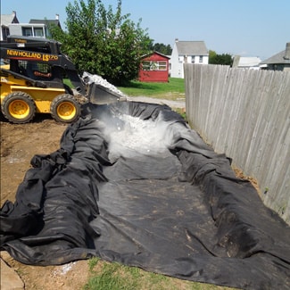 A small construction vehicle from Steve L. Dellinger Contractor is pouring gravel into a large rectangular pit lined with black geotextile fabric in a backyard. There are houses and trees in the background, and a wooden fence runs along one side of the pit, showcasing their excavating expertise for both residential and commercial projects.