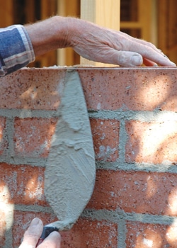 A close-up photo of a person's hands using a trowel to apply mortar between bricks in a wall. One hand holds the trowel while the other steadies the brickwork. The image captures Steve L. Dellinger Contractor at work, showcasing the construction process of bricklaying for residential and commercial projects.