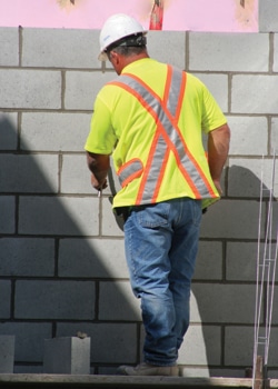 A construction worker in a bright yellow shirt with a reflective orange vest and a white hard hat is building a wall with cement blocks. The worker, engaged in residential and commercial projects, is facing the wall and appears to be applying or adjusting the blocks.