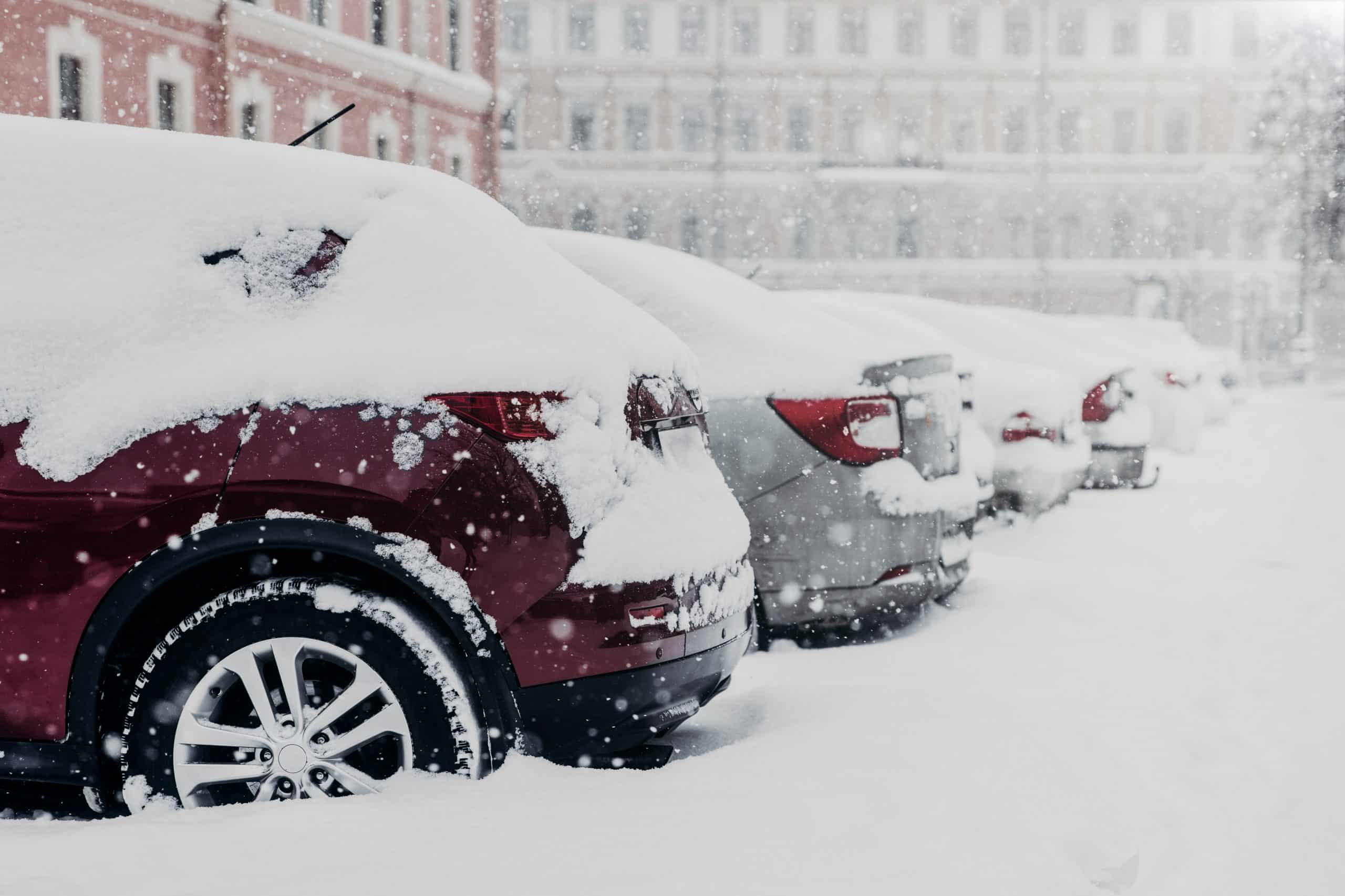 A row of parked cars covered in thick snow during a snowfall. The vehicles, lined up next to a building, are mostly unrecognizable due to the heavy snow accumulation on the flat concrete surface. Snowflakes are visible in the air, creating a wintry atmosphere.