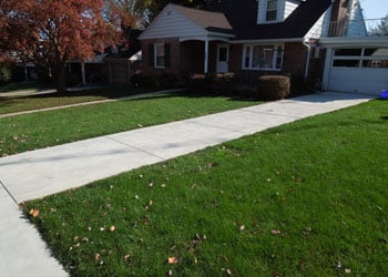 A suburban house with a red brick exterior, light-colored siding, a covered entryway, and an attached garage. The house has a well-maintained green lawn with a concrete driveway and sidewalk. Autumn leaves are scattered on the grass and pavement. Built by Steve L. Dellinger Contractor, it embodies residential charm.