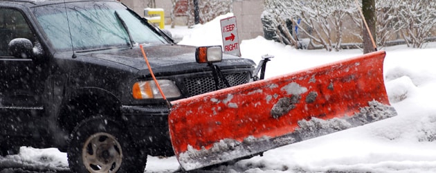 A black truck with a red snowplow attached to its front, labeled "Steve L. Dellinger Contractor," clears snow from a snowy street. Snow is falling and the surroundings are covered in snow. A sign in the background reads "KEEP RIGHT.