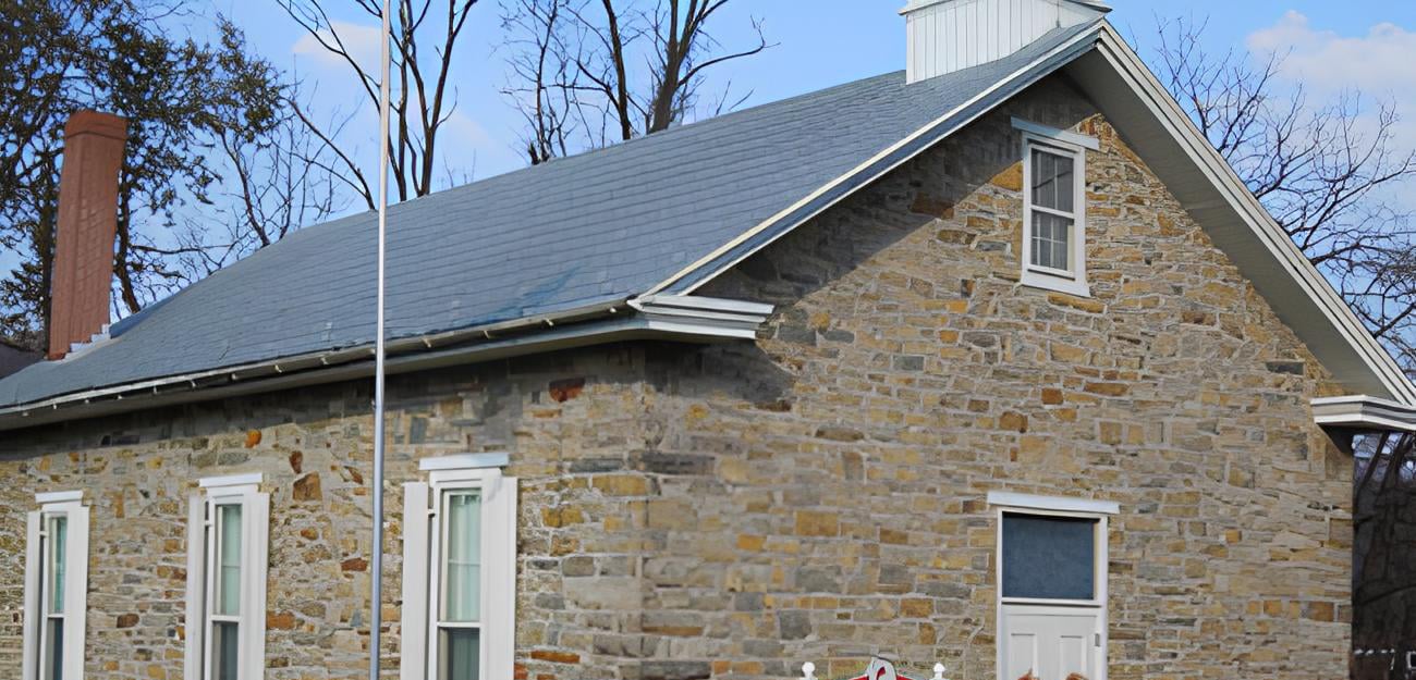 A rustic stone house with a steep gray roof and white-trimmed windows and doors, complemented by a red brick chimney. The home is surrounded by leafless trees under a blue sky, with an American flagpole standing proudly beside it. Steve L. Dellinger Contractor specializes in both residential and commercial projects like this.