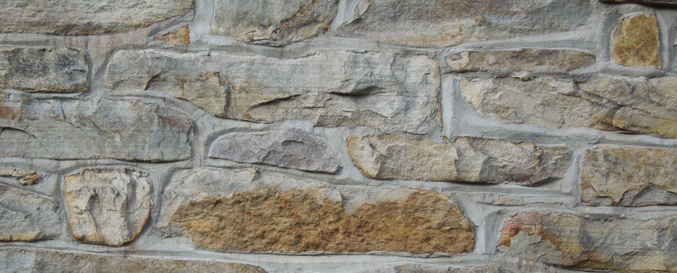 A close-up view of a stone wall made of large, irregularly-shaped stones mortared together. The stones vary in color, including shades of gray, beige, and brown, creating a rugged and textured surface often seen in both residential and commercial settings.