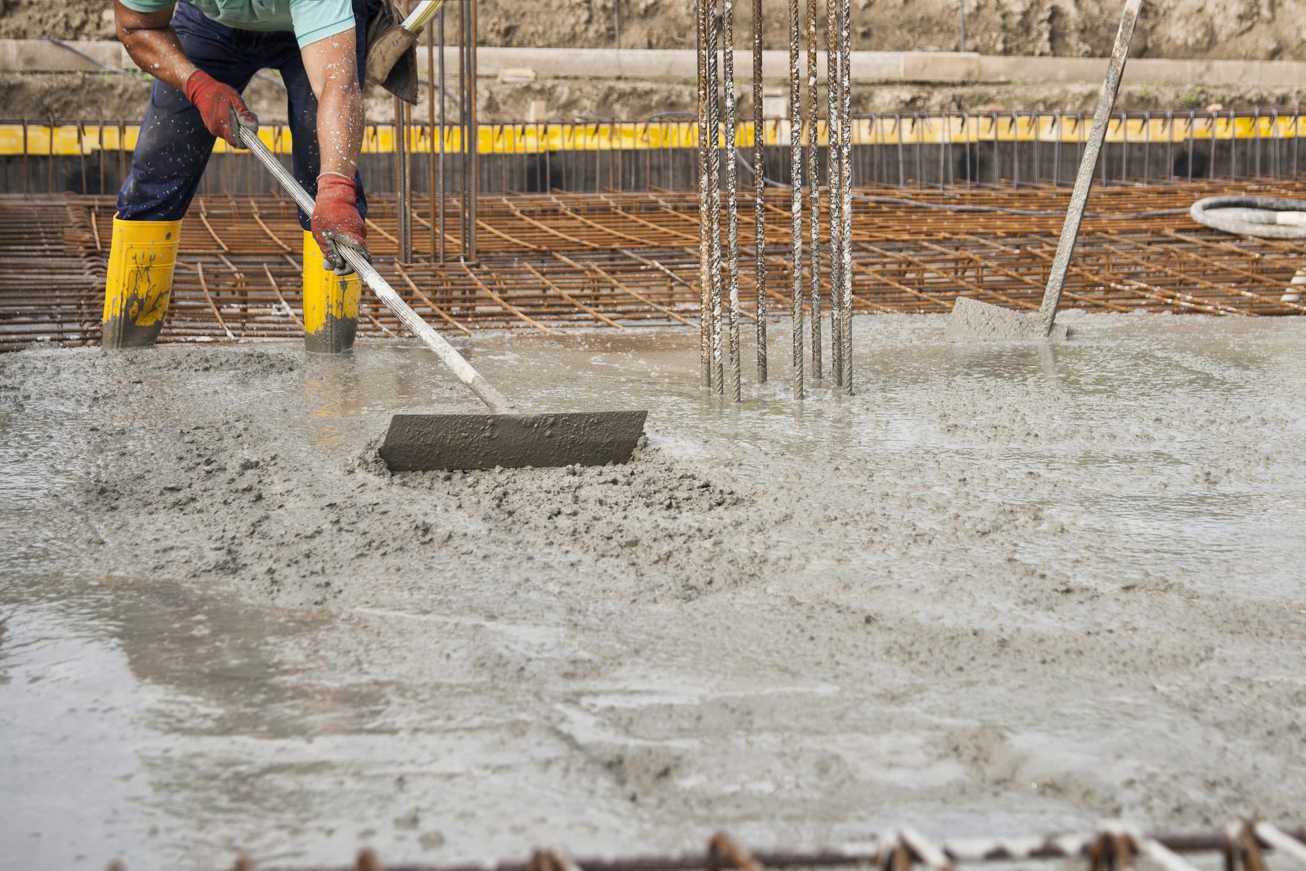 A construction worker wearing a green shirt, yellow safety boots, and red gloves spreads wet concrete at a building site using a long-handled tool. Rebar can be seen protruding from the concrete around the worker. Steve L. Dellinger Contractor specializes in residential and commercial projects.