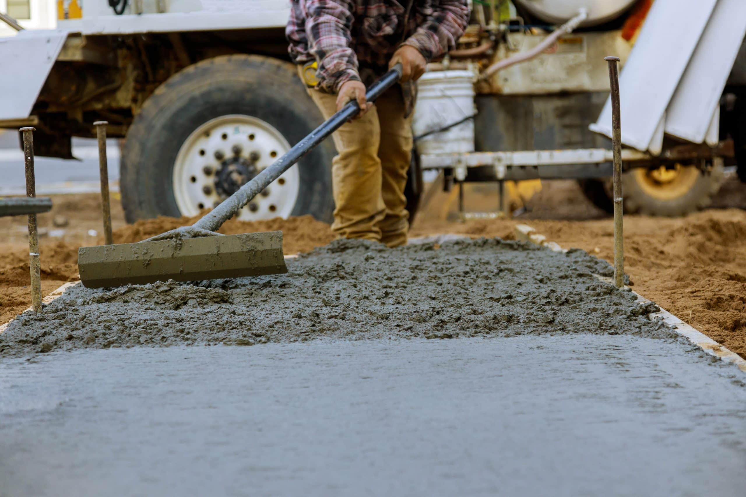 A construction worker in a checkered shirt smoothens wet concrete on a ground surface using a long tool. A truck with large wheels is visible in the background, and the surrounding area appears sandy. Metal stakes are placed along the edges of the concrete path for this Steve L. Dellinger Contractor project.